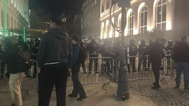 Oameni adunați seara în spatele gardurilor metalice, în fața unei clădiri luminate