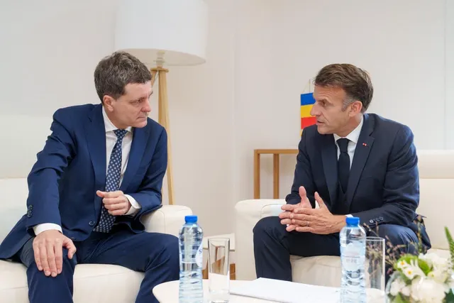 Doi bărbați în costume discutând, așezați în fotolii albe, cu drapelul României în spate
