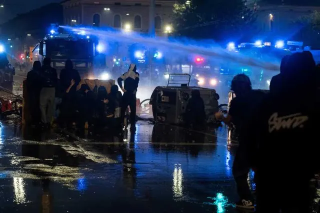 Tun cu apă al poliției în acțiune noaptea, cu girofaruri albastre și protestatari pe o stradă udă