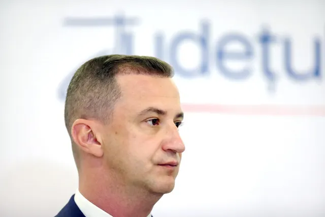 Bărbat din profil, în costum, în fața unui panou alb cu litere albastre