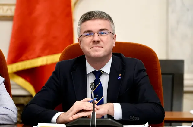 Bărbat cu ochelari, în costum și cravată, așezat la un pupitru cu microfon, cu drapele în spate