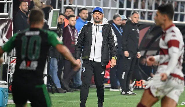 Antrenor în geacă albă și șapcă albastră pe marginea terenului, între fotbaliști în echipamente verde și alb-vișiniu