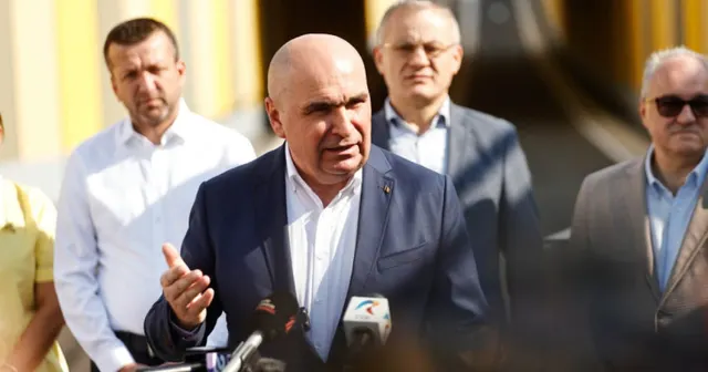 Bărbat chel, în costum închis, vorbind la microfoanele presei, înconjurat de alți bărbați