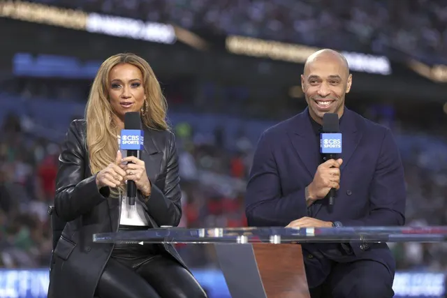 Doi prezentatori TV, o femeie blondă și un bărbat în costum închis, cu microfoane, la un pupitru pe stadion