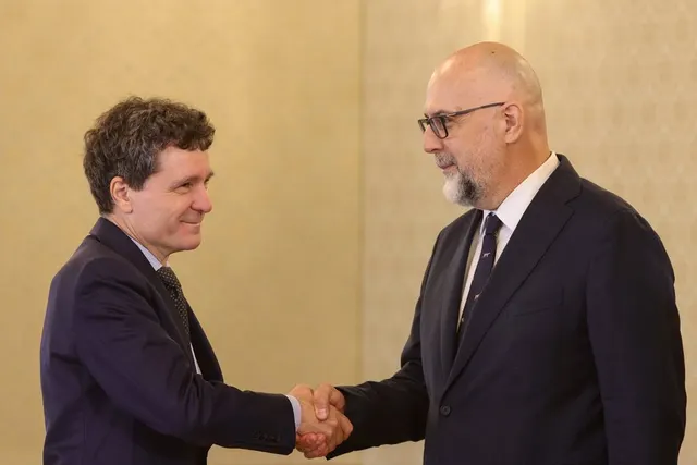 Doi bărbați în costume își strâng mâna într-o sală cu pereți crem