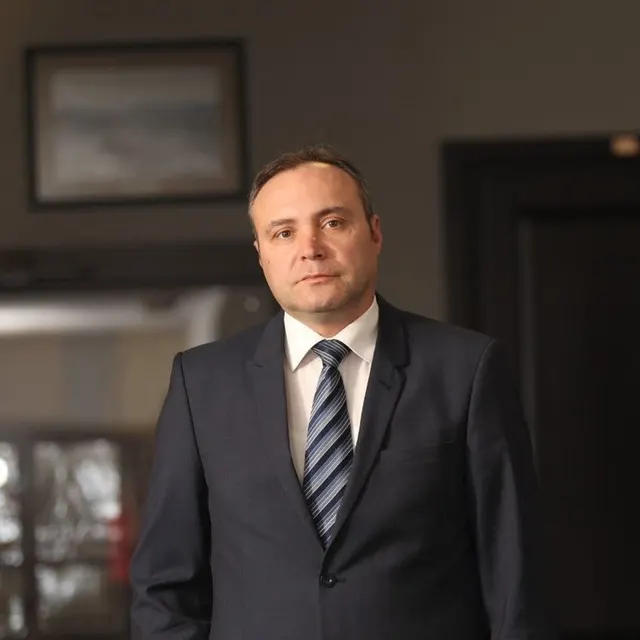 Bărbat în costum închis și cravată în dungi, în picioare într-un birou cu un tablou pe perete