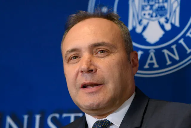 Bărbat vorbind în fața unui panou albastru cu stema României și inscripția unei instituții