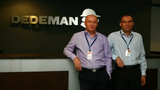 Frații Pavăl, proprietarii Dedeman, în picioare în fața siglei companiei de pe un perete