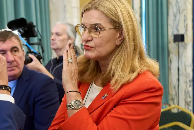 Femeie cu ochelari, în sacou roșu, cu mâinile împreunate la bărbie, lângă un bărbat și o cameră de filmat
