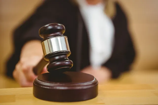Ciocănel de judecător din lemn pe suportul lui, cu un magistrat în robă neagră în fundal
