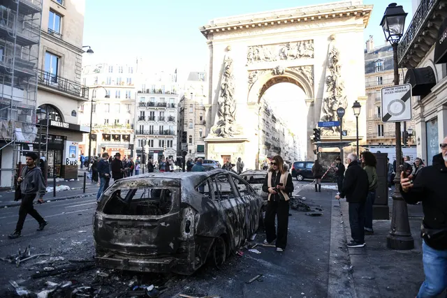Mașină incendiată pe o stradă din Paris, cu un arc de triumf în fundal și trecători care privesc pagubele
