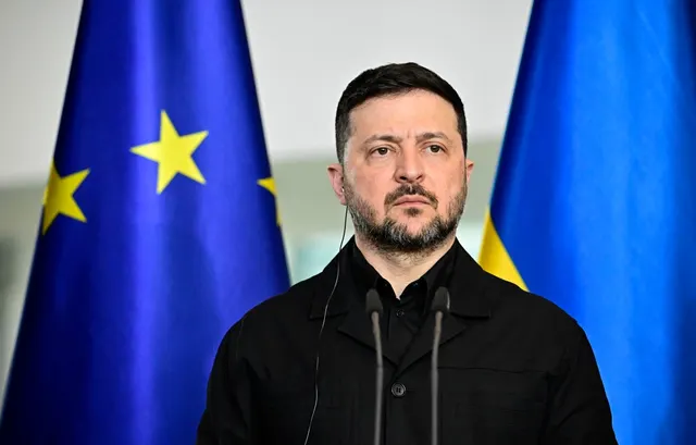 Volodimir Zelenski, îmbrăcat în negru, la un pupitru cu microfoane, între drapelele UE și Ucrainei