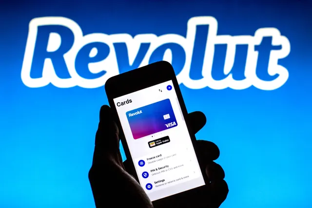 Mână care ține un telefon cu aplicația Revolut deschisă, în fața siglei Revolut pe fundal albastru